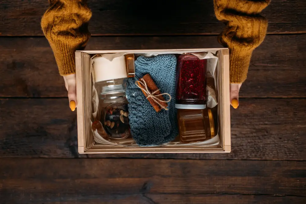 An overhead view of hands holding a wooden gift crate packed with a candle, a knitted throw, jam and honey