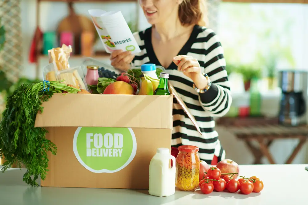 Someone reading a recipe card beside a Food Delivery box packed with fresh produce