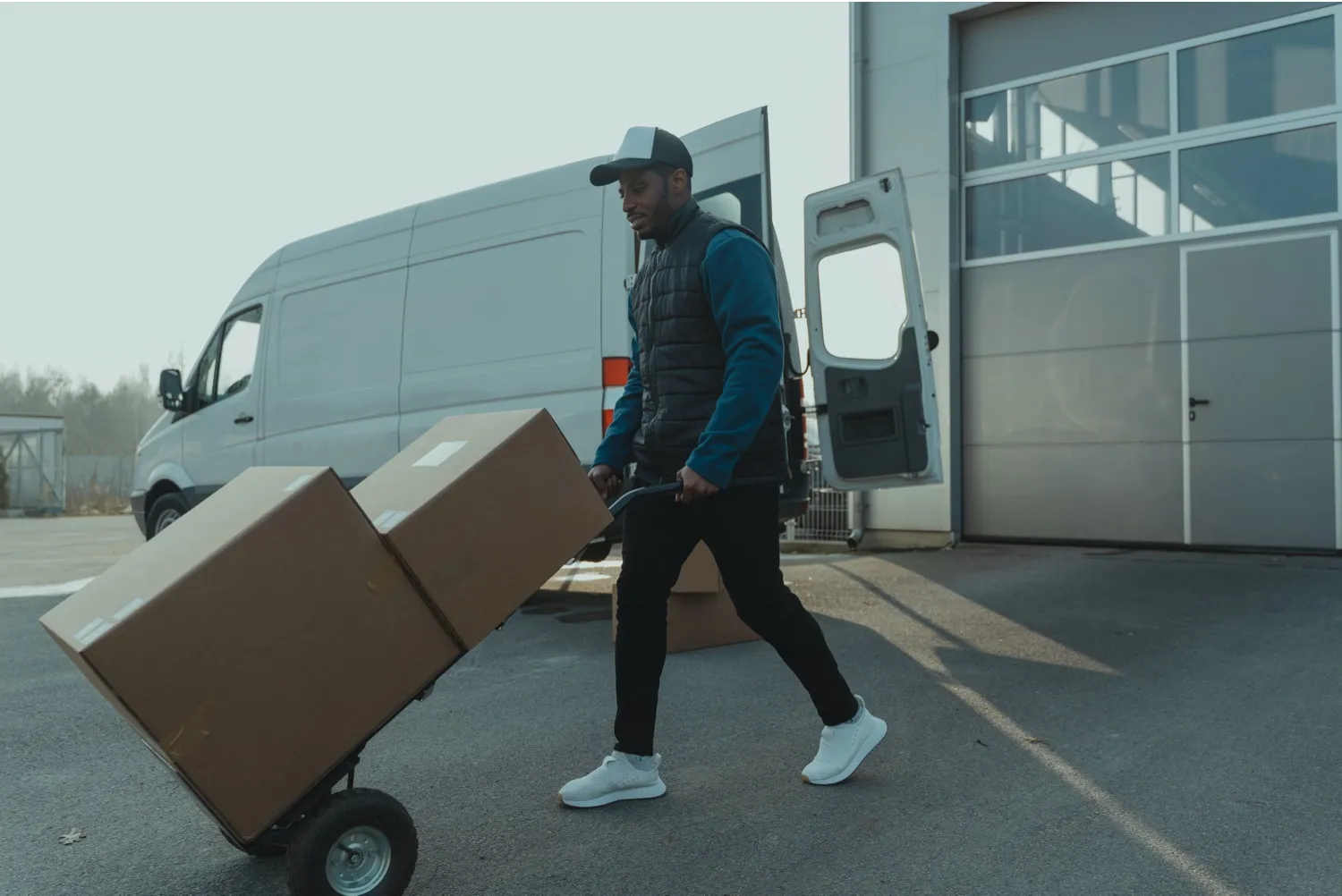 A courier wheeling two large cartons on a sack truck past an open van at a depot