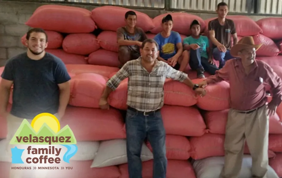 The Velasquez Family Coffee team smiling in front of a wall of stacked coffee sacks, with the company's logo in the corner