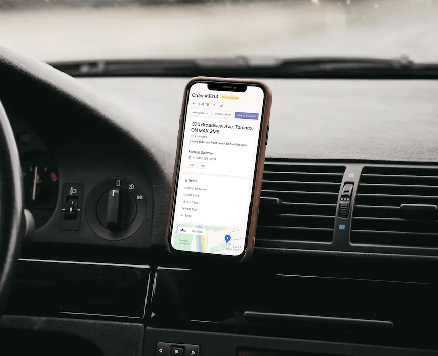 A phone mounted on a car dashboard vent showing Delivery View for one order, with the address, delivery note, Call and Text buttons, items and a map