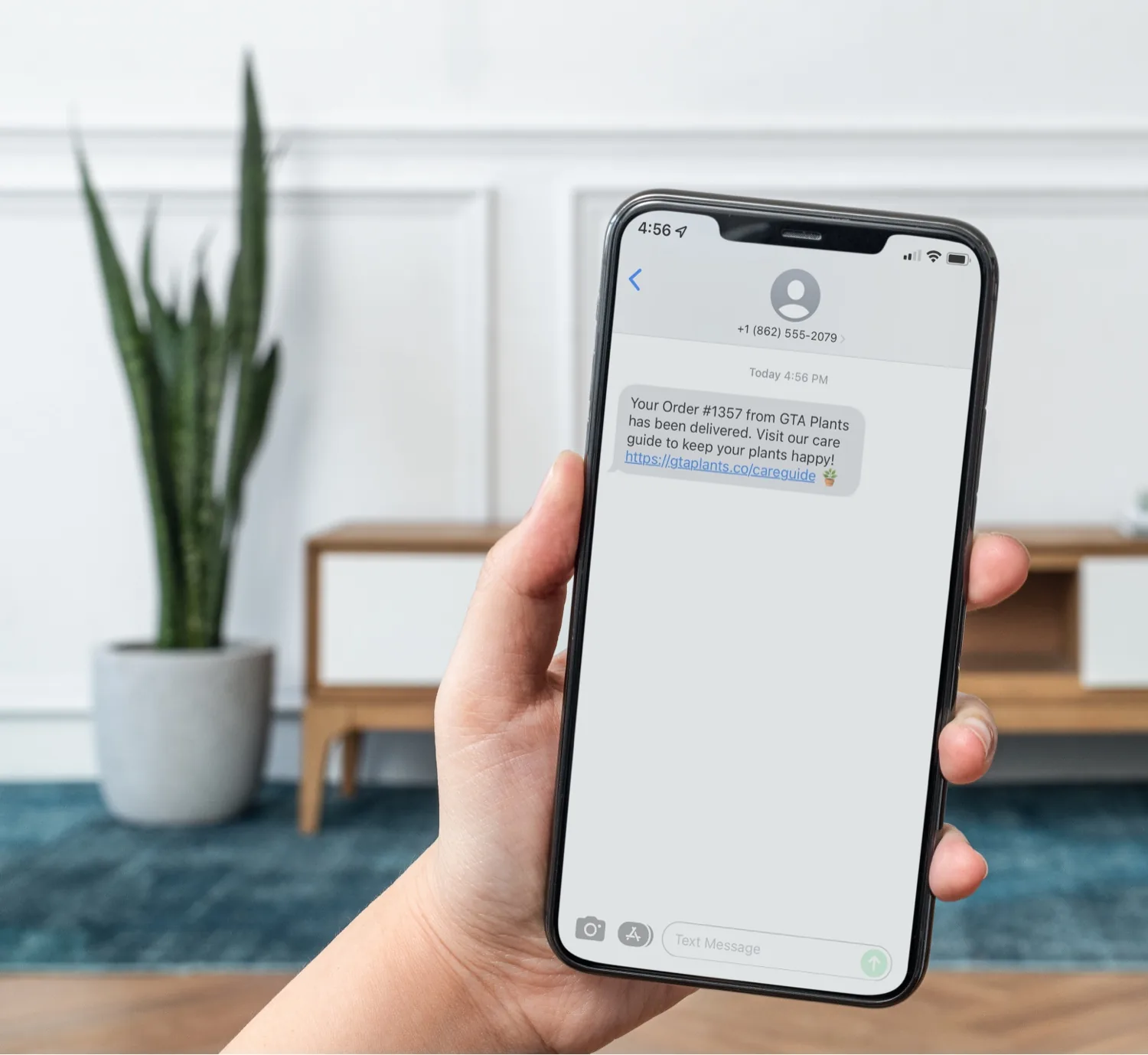 A hand holding a phone showing a text from GTA Plants saying order #1357 has been delivered, with a link to the shop’s plant care guide