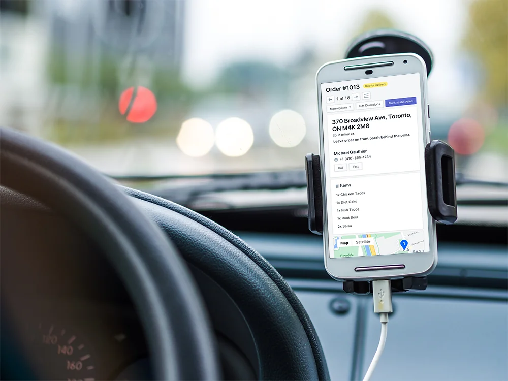 A phone in a dashboard mount showing Driver View for an order out for delivery, with its address, delivery note and items